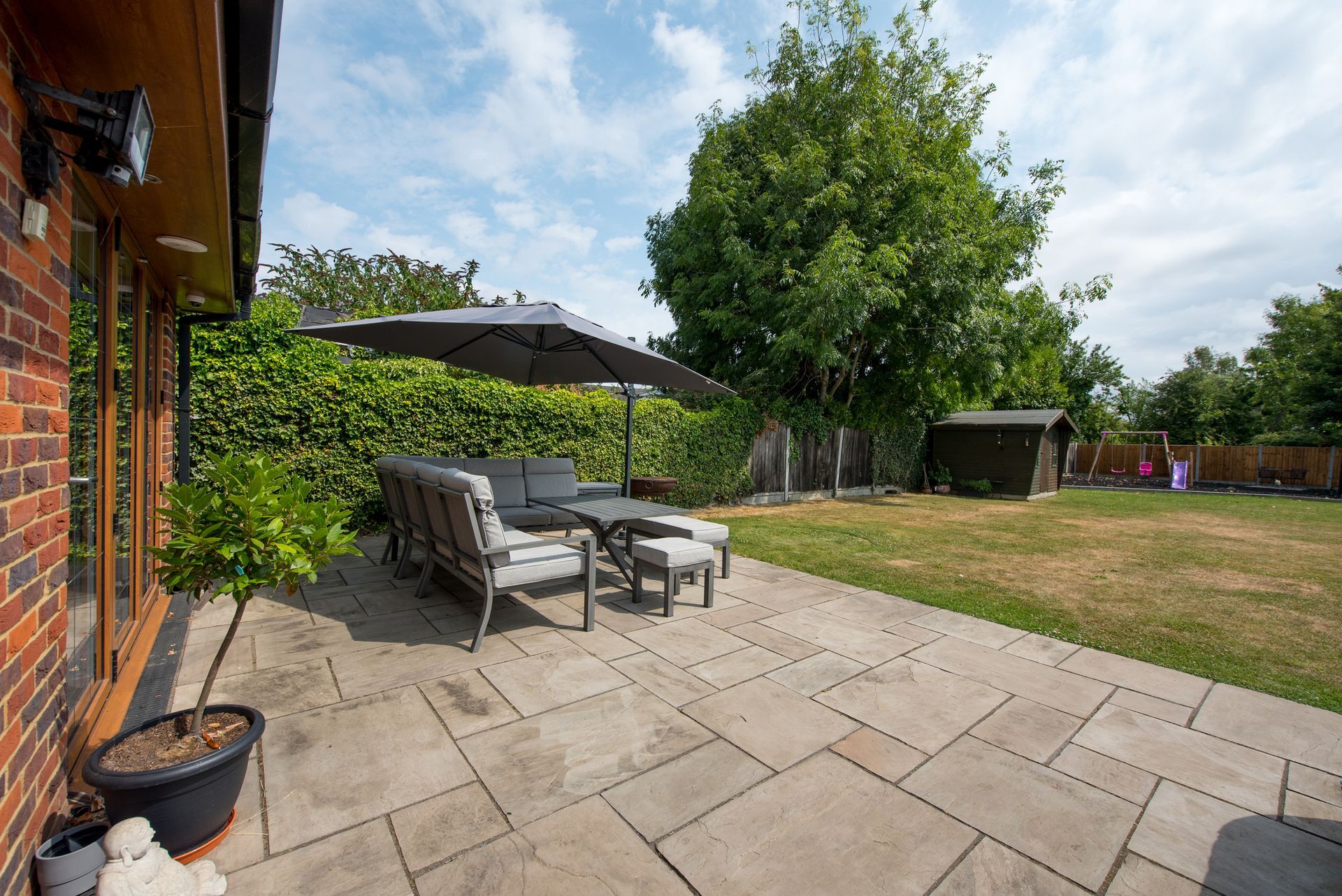 Patio with seating under an umbrella, brick house, green lawn, shed, and trees under a sunny sky.