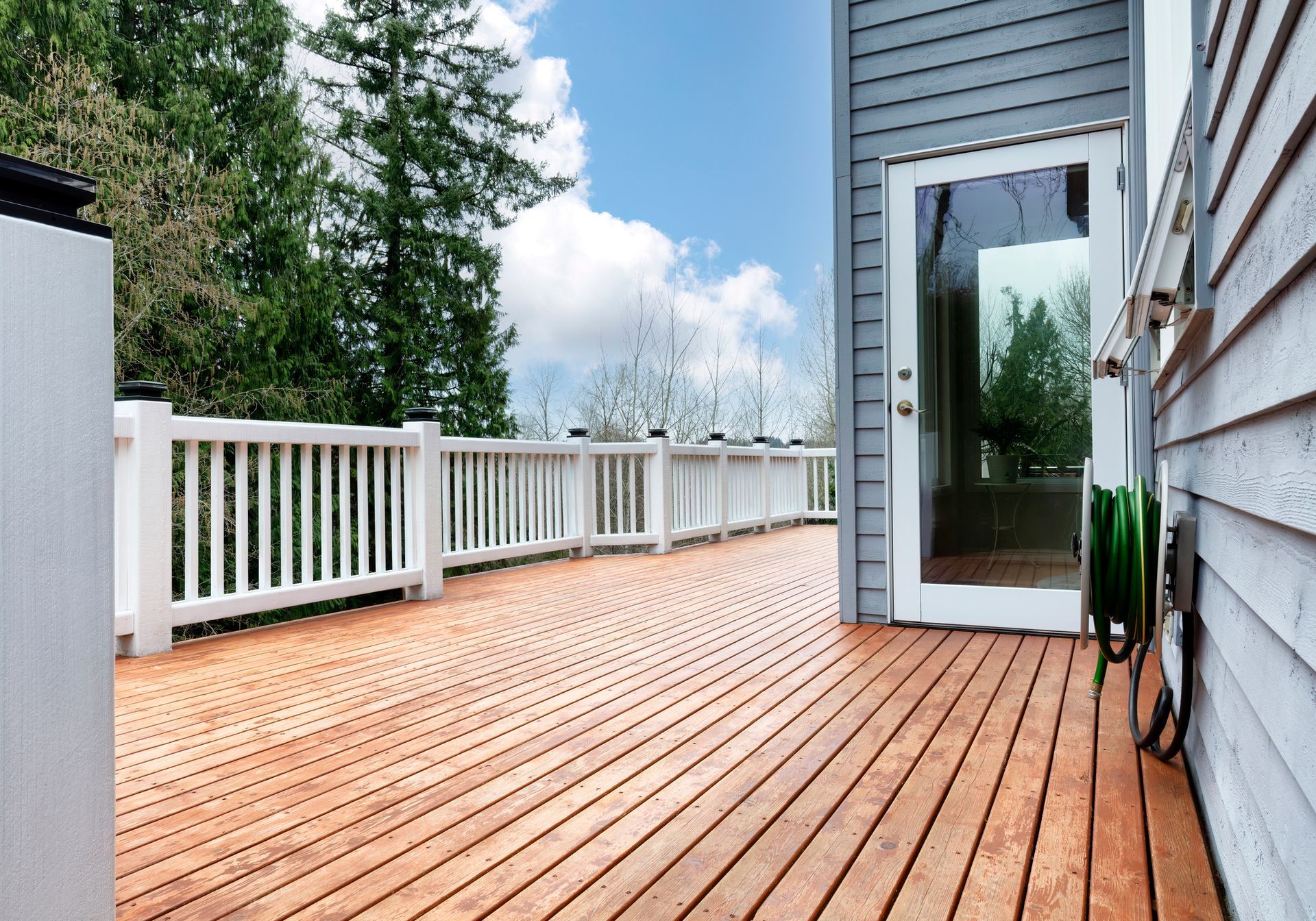 Wooden deck with white railing, glass door, and blue siding. Hose attached. Sunny day, trees in background.