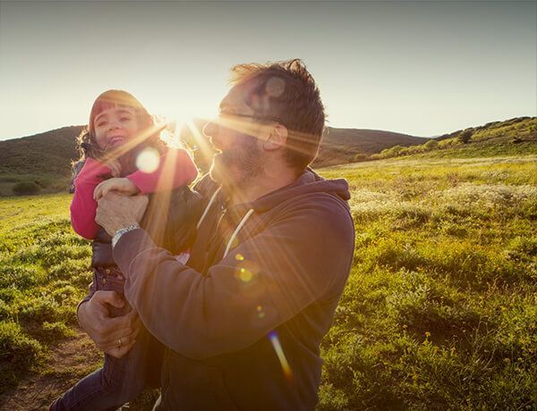 Adult holding a smiling child in a sunlit grassy field with hills in the background