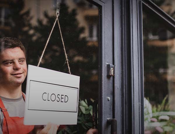 Storefront sign reading “CLOSED” hanging in a glass door as a worker stands nearby.