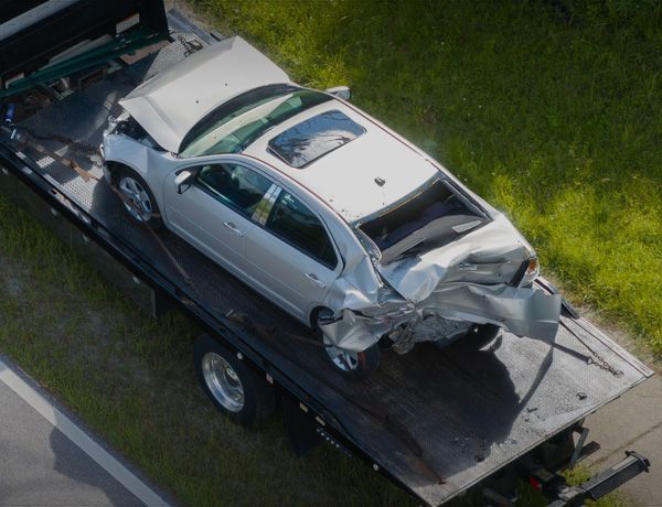 Wrecked white car loaded on a tow truck, with severe front-end damage and a crushed roof.