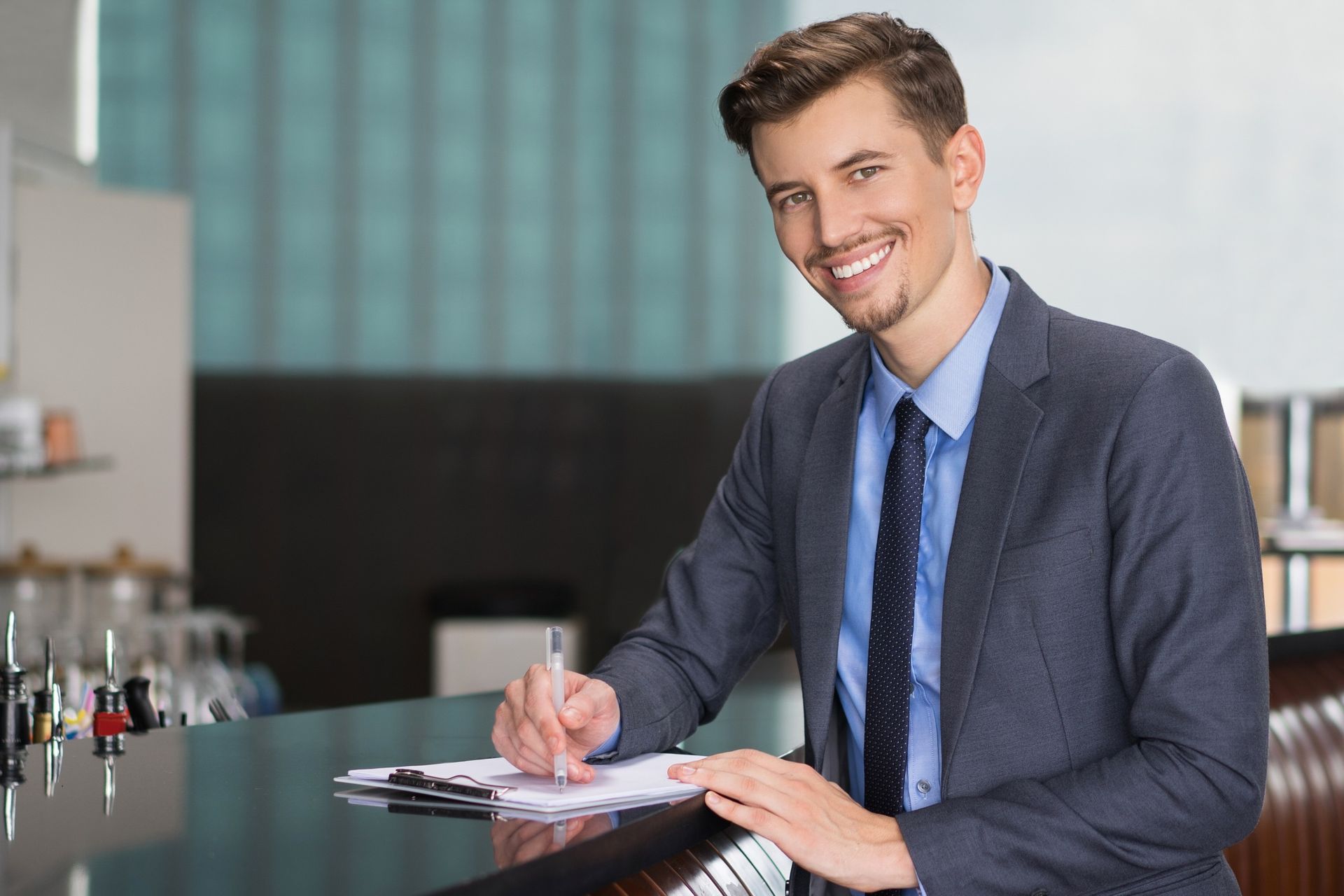 Man in a blue suit smiling while writing on a clipboard at a bar counter