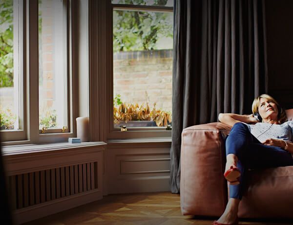 Woman reclining on a sofa in a bright living room beside large windows and dark curtains