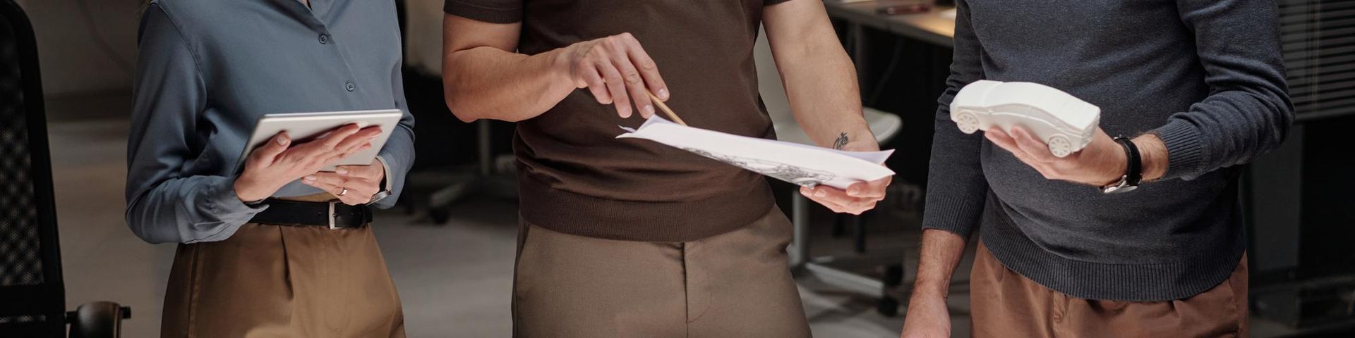 Three people standing indoors, holding notebooks and papers while discussing.