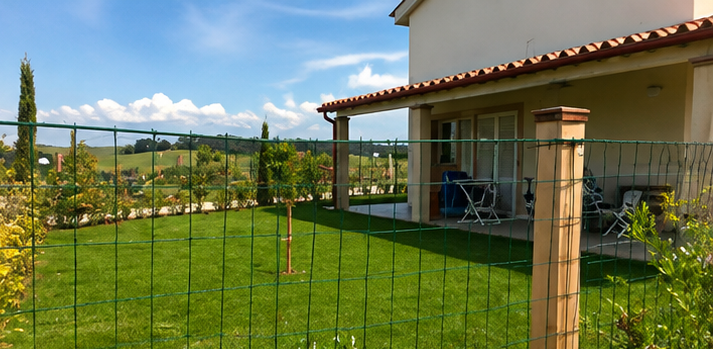 Un prato verde accanto a una casa con un cortile recintato e un giardino sotto un cielo azzurro.