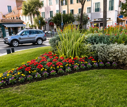 Un'aiuola colorata in una piazza cittadina con auto parcheggiate e edifici color pastello sullo sfondo.