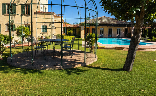 Cortile con giardino, gazebo in metallo, prato verde, alberi e una piscina blu accanto a un edificio.