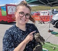 Smiling person in glasses holding a bird at an outdoor fair with red buses and tents in the background