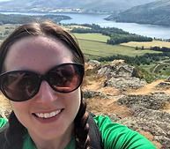 Smiling person in sunglasses on a rocky overlook above a lake and green valley