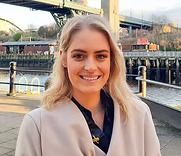 Smiling woman in a beige coat standing by a waterfront with boats and a bridge in the background