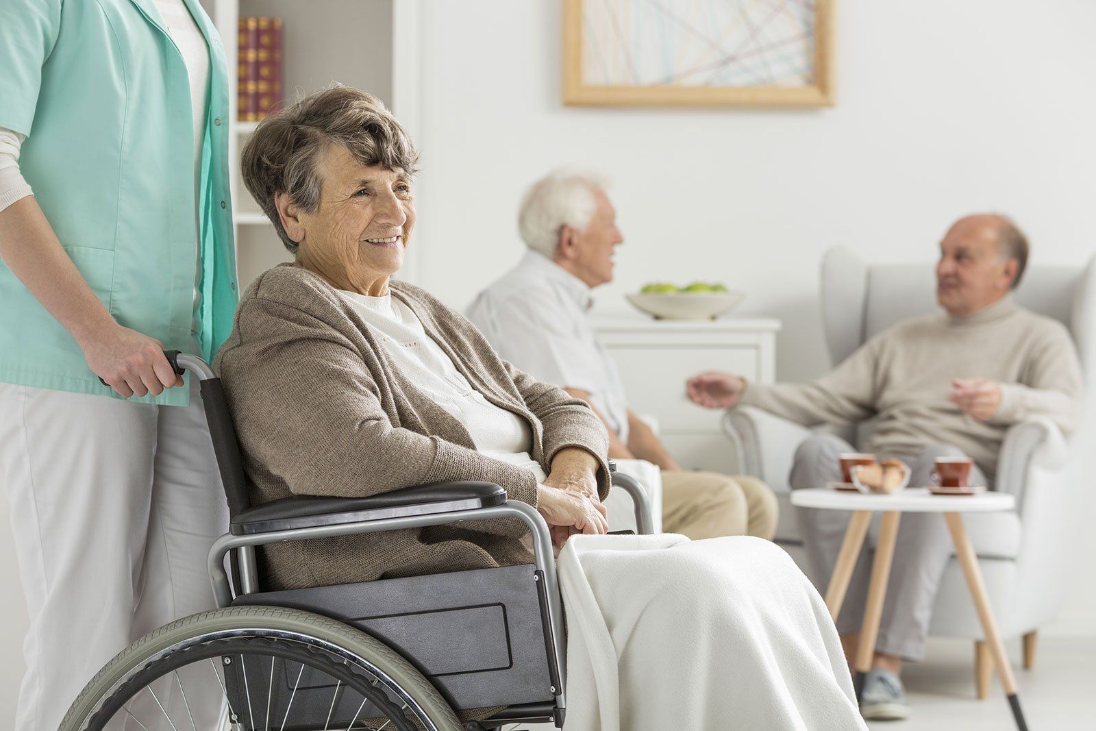 Donna sorridente su sedia a rotelle, assistita da un assistente in un luminoso soggiorno insieme ad altri residenti.