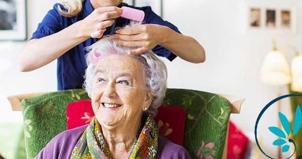 Una donna sorride mentre un'altra le mette un bigodino tra i capelli argentati, in casa.