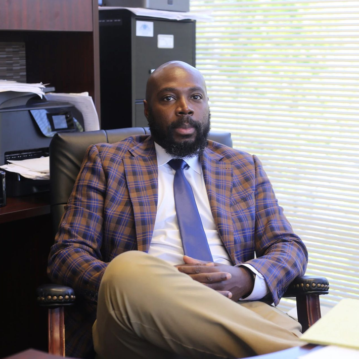Man seated in an office chair, wearing a plaid blazer and tie, with a desk and filing cabinets behind him.