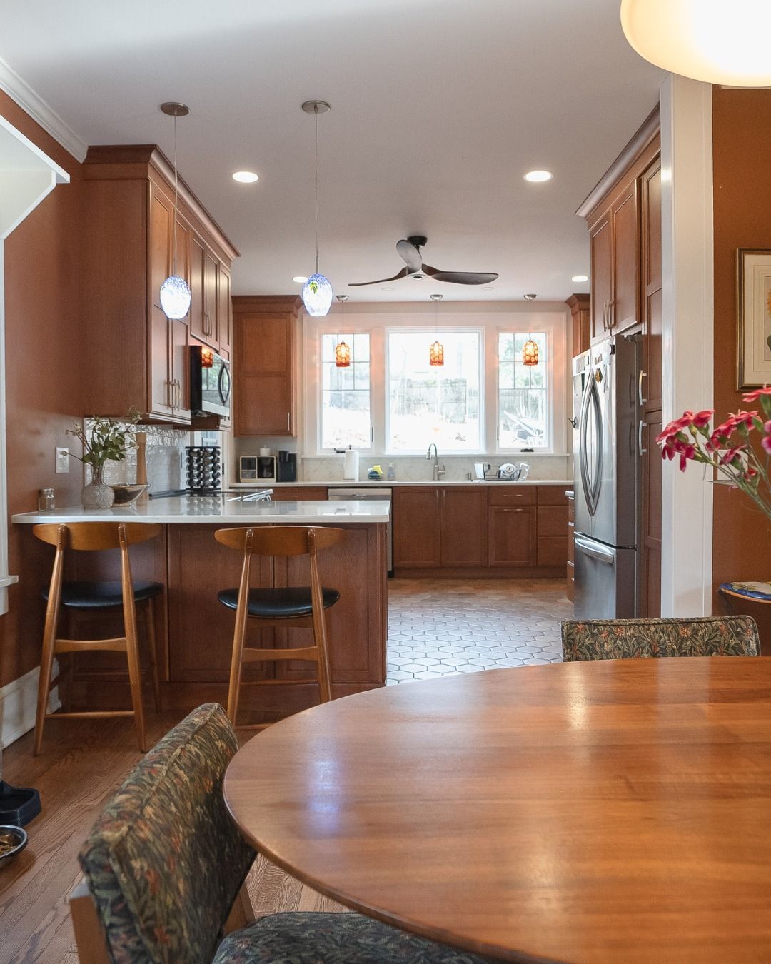 Warm kitchen with wood cabinets, island stools, round dining table, and large windows over a patterned floor.