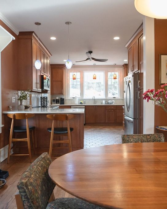 Warm kitchen with wood cabinets, island stools, round dining table, and large windows over a patterned floor.