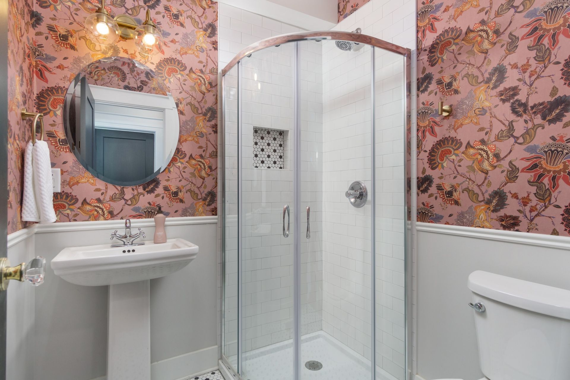 Small bathroom with floral wallpaper, pedestal sink, shower stall, and toilet.