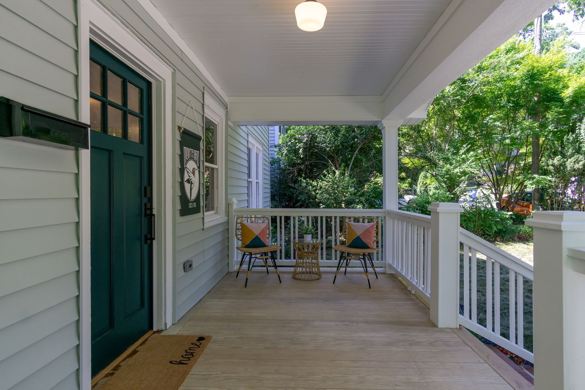 Front porch with white railings, green door, two chairs, and leafy trees beyond.