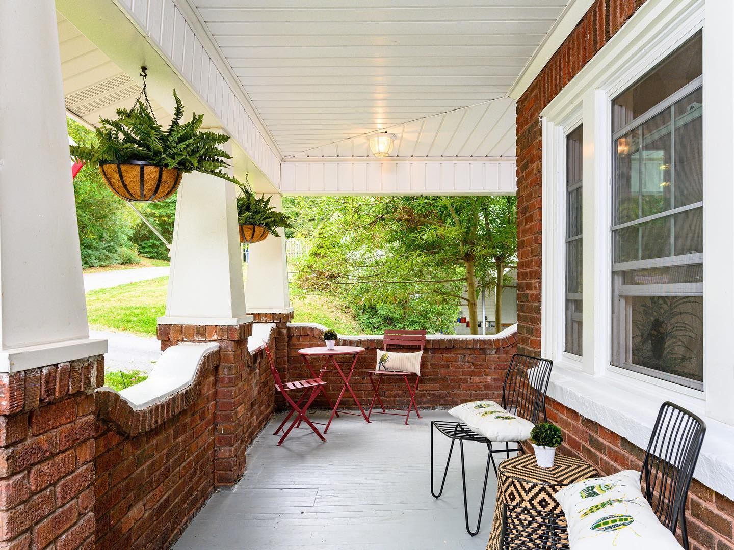 Covered front porch with brick railing, hanging plants, and small tables and chairs overlooking a green yard