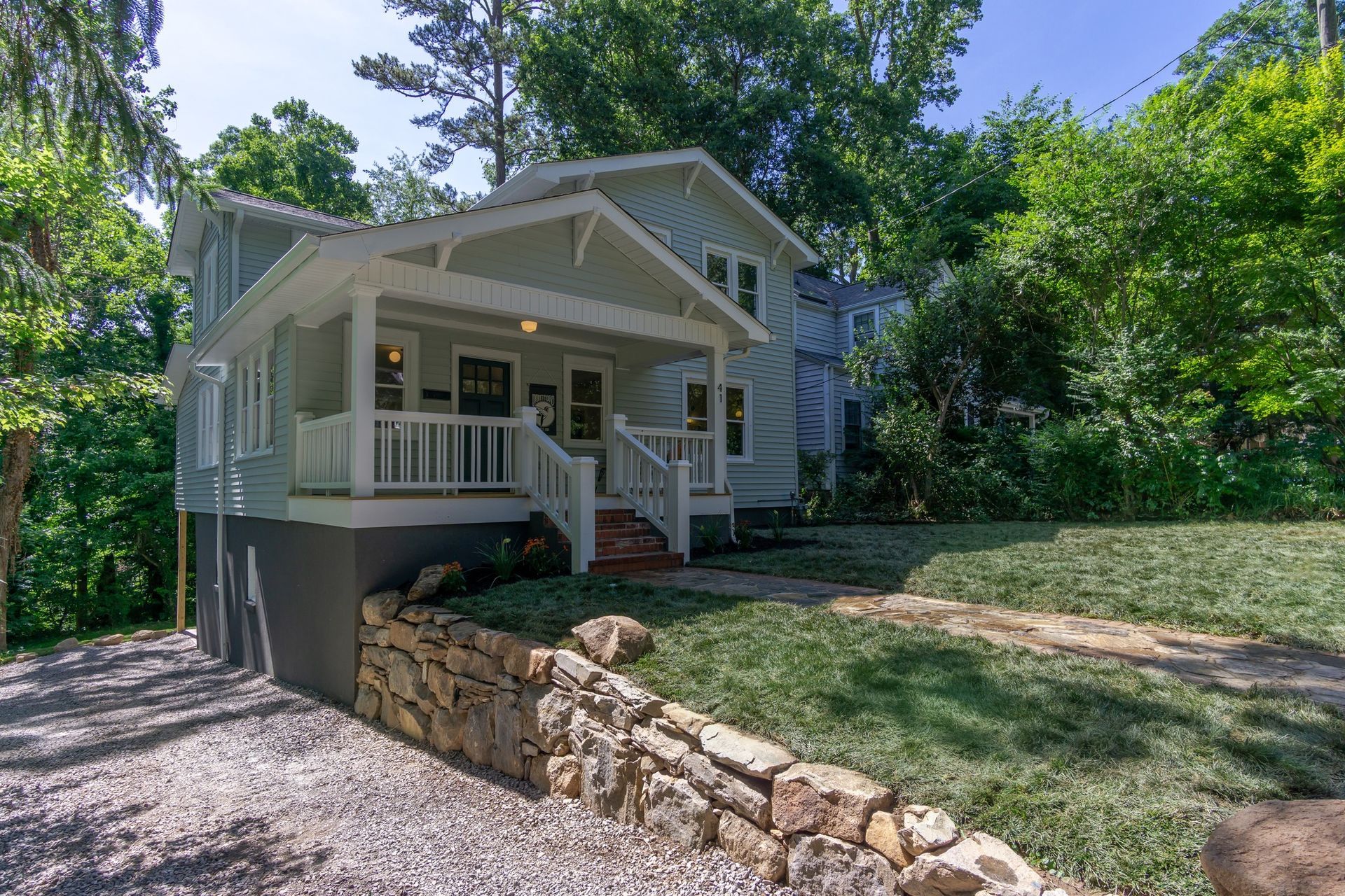 White house with front porch and stone retaining wall on a sloped, tree-lined lot