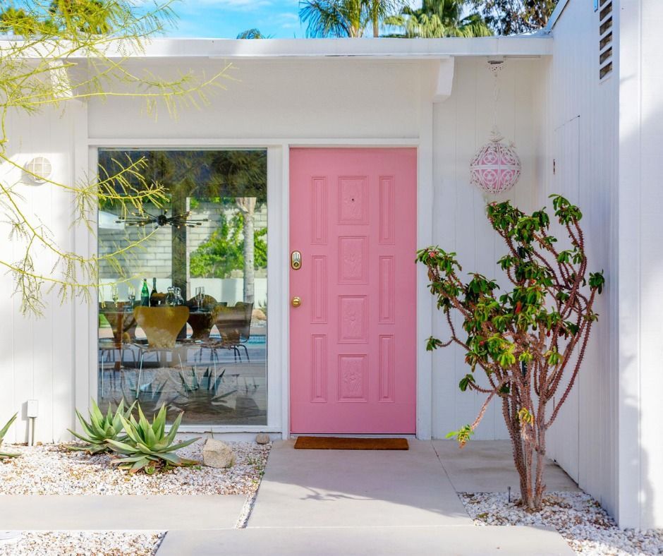 Modern white house entry with a pink front door, glass window, and desert plants in bright sunlight