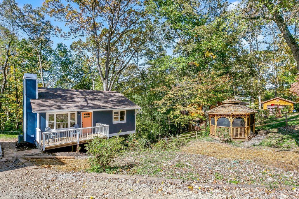 Small blue house with porch in a wooded yard beside a wooden gazebo