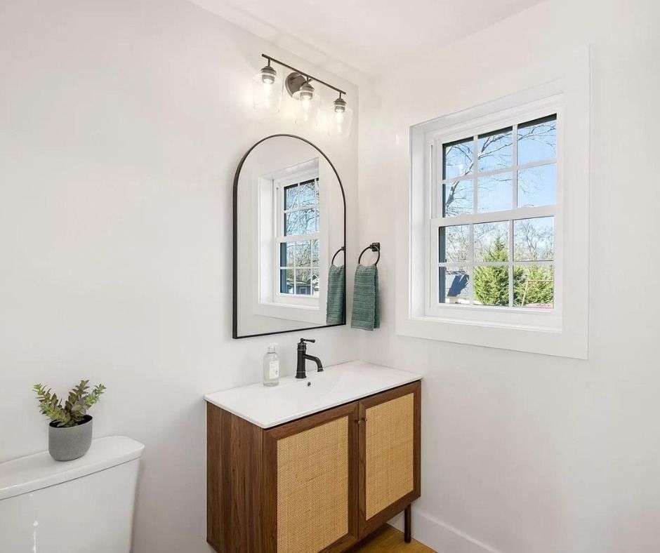 Small white bathroom with a wooden vanity, mirror, and window letting in daylight.