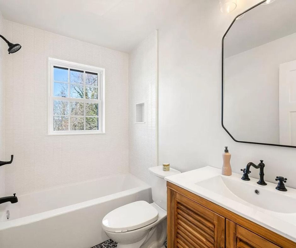 Bright white bathroom with black fixtures, wooden vanity, bathtub, toilet, and window near mirror