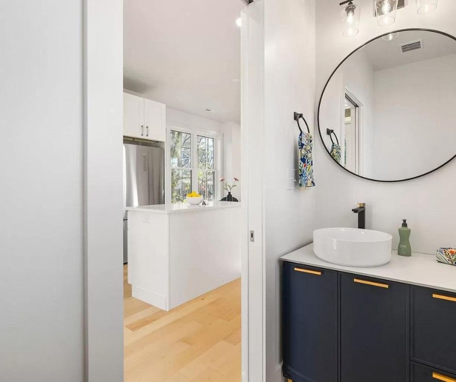 Modern bathroom with navy vanity, round mirror, and a view into a bright kitchen beyond.