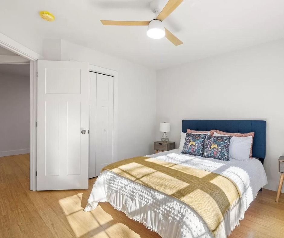 Bright bedroom with white walls, wood floor, blue headboard bed, and ceiling fan.