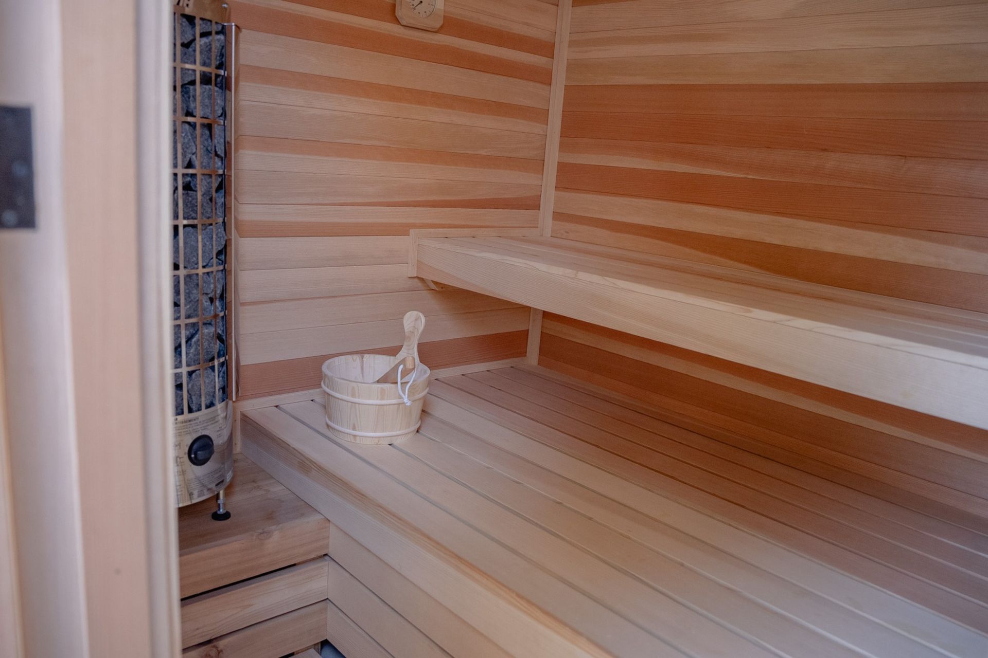 Wood-paneled sauna with wooden benches and a bucket on the lower seat, lit by warm light