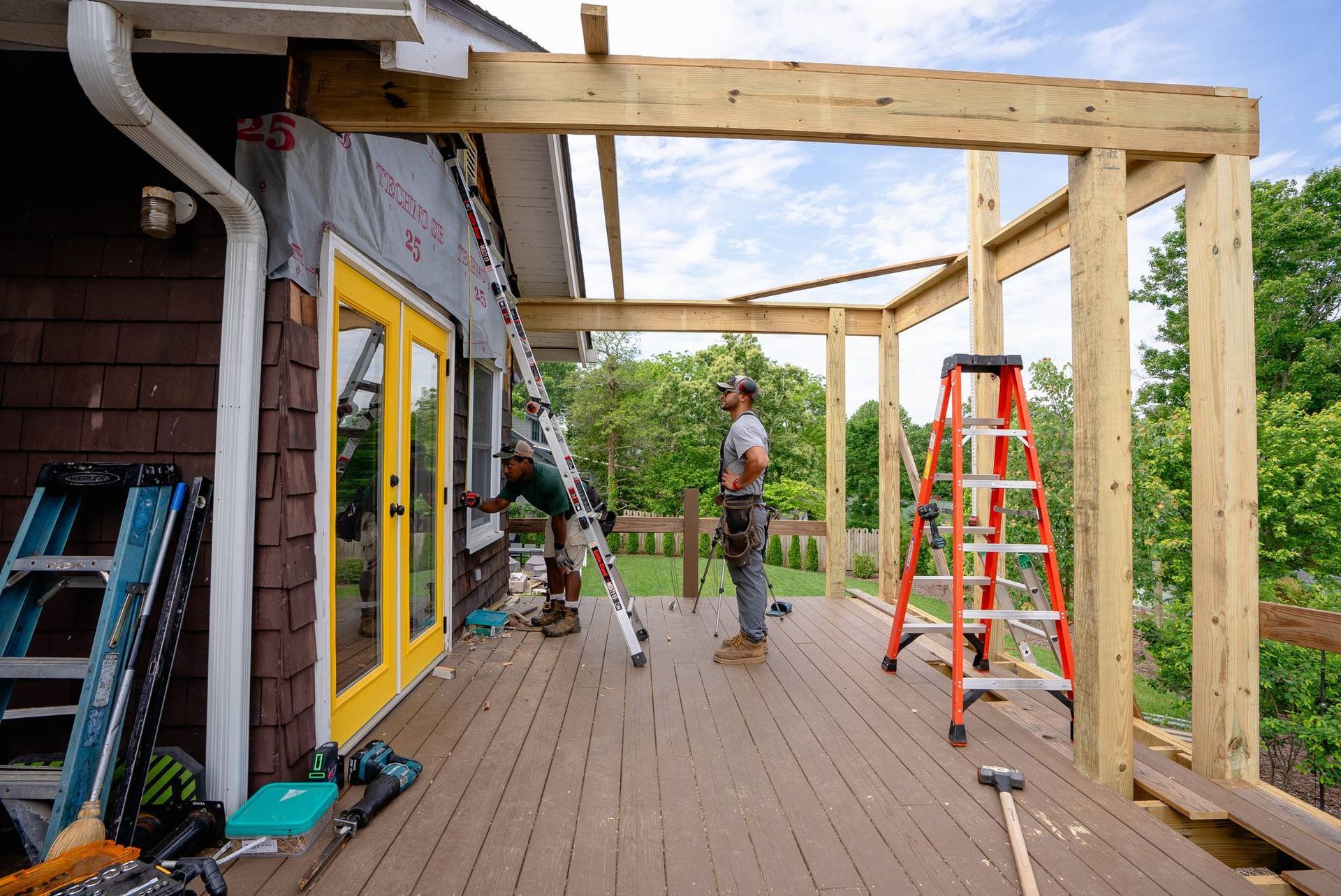 Workers building a wooden patio pergola beside a house, with ladders and tools on deck.