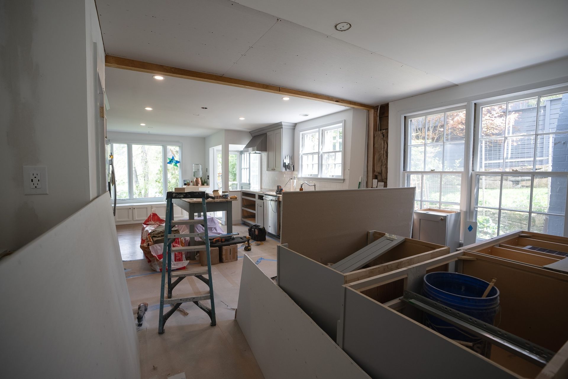 Renovation site with drywall, tools, and a ladder in a bright room with large windows