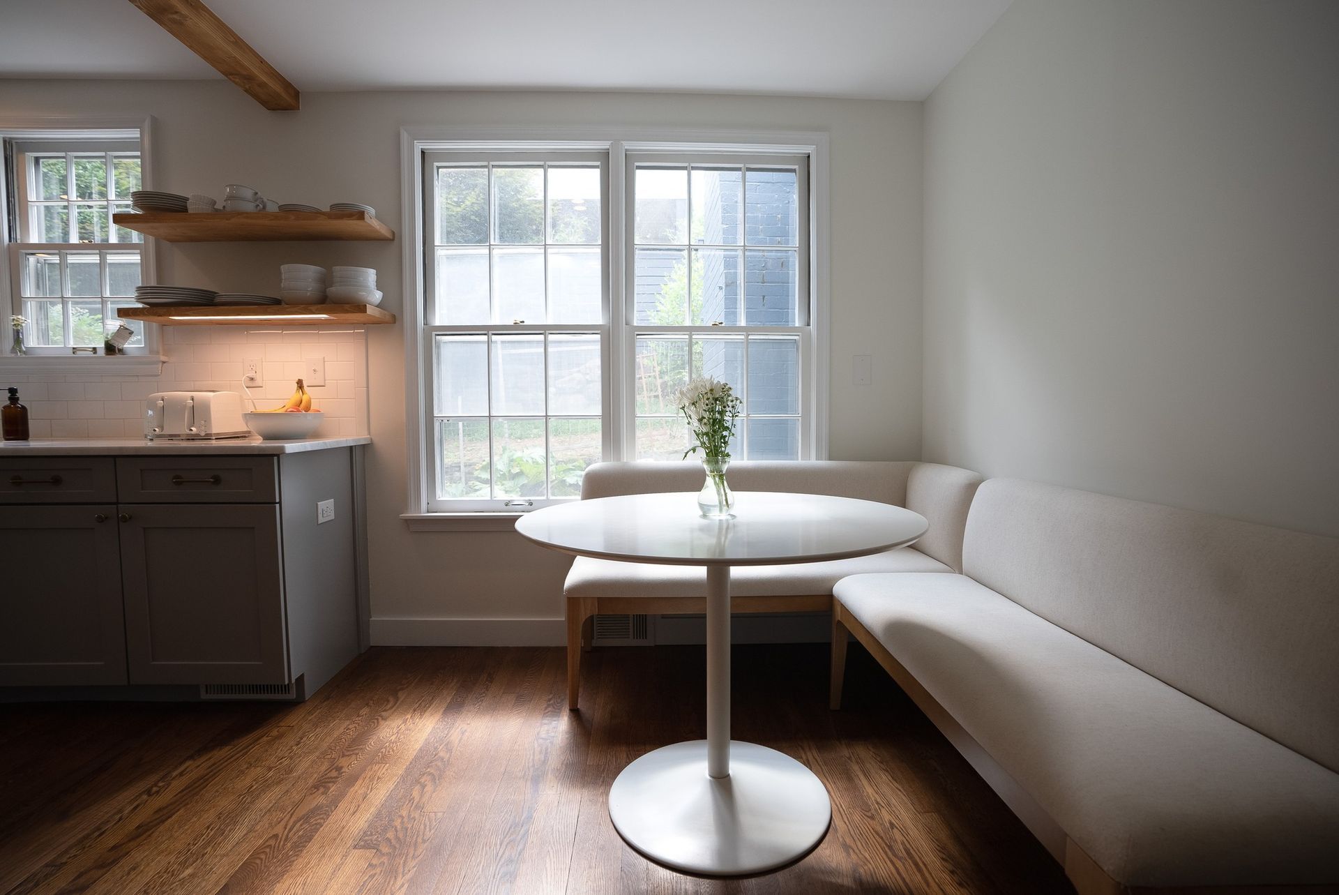 Bright living room with white sofa, round table, window light, and kitchen nook with shelves and sink.
