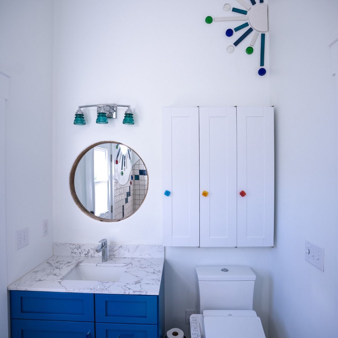 Bright white bathroom with blue vanity, round mirror, wall cabinet, toilet, and towel rack