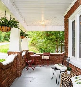 Covered brick porch with hanging plants, bistro table, chairs, and cushioned bench overlooking greenery