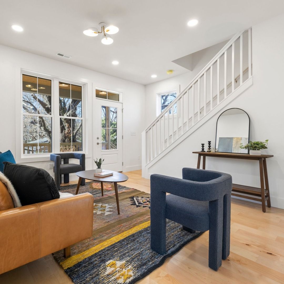 Bright living room with blue chairs, tan sofa, stairs, and a wooden console table by the window