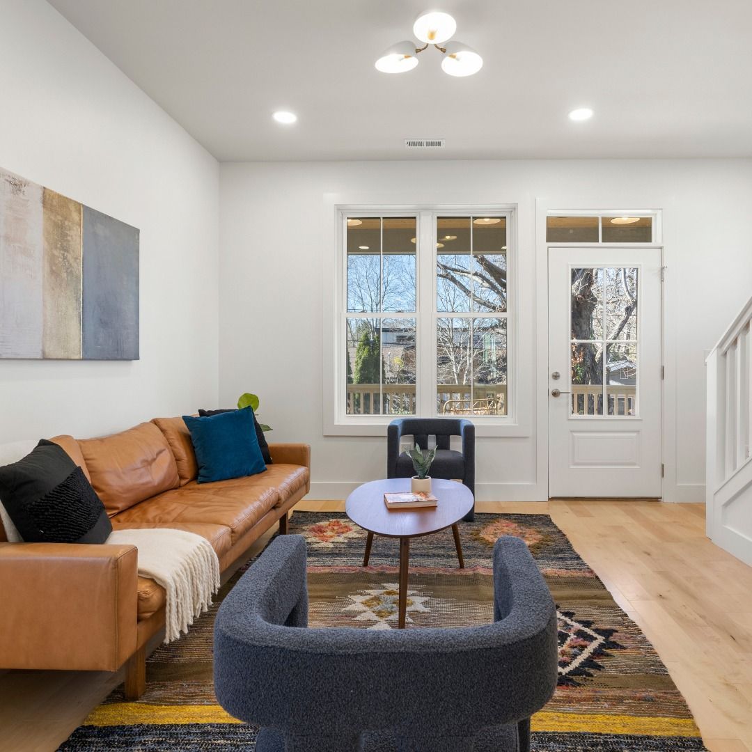 Bright living room with tan sofa, dark chair, round coffee table, patterned rug, and white front door.