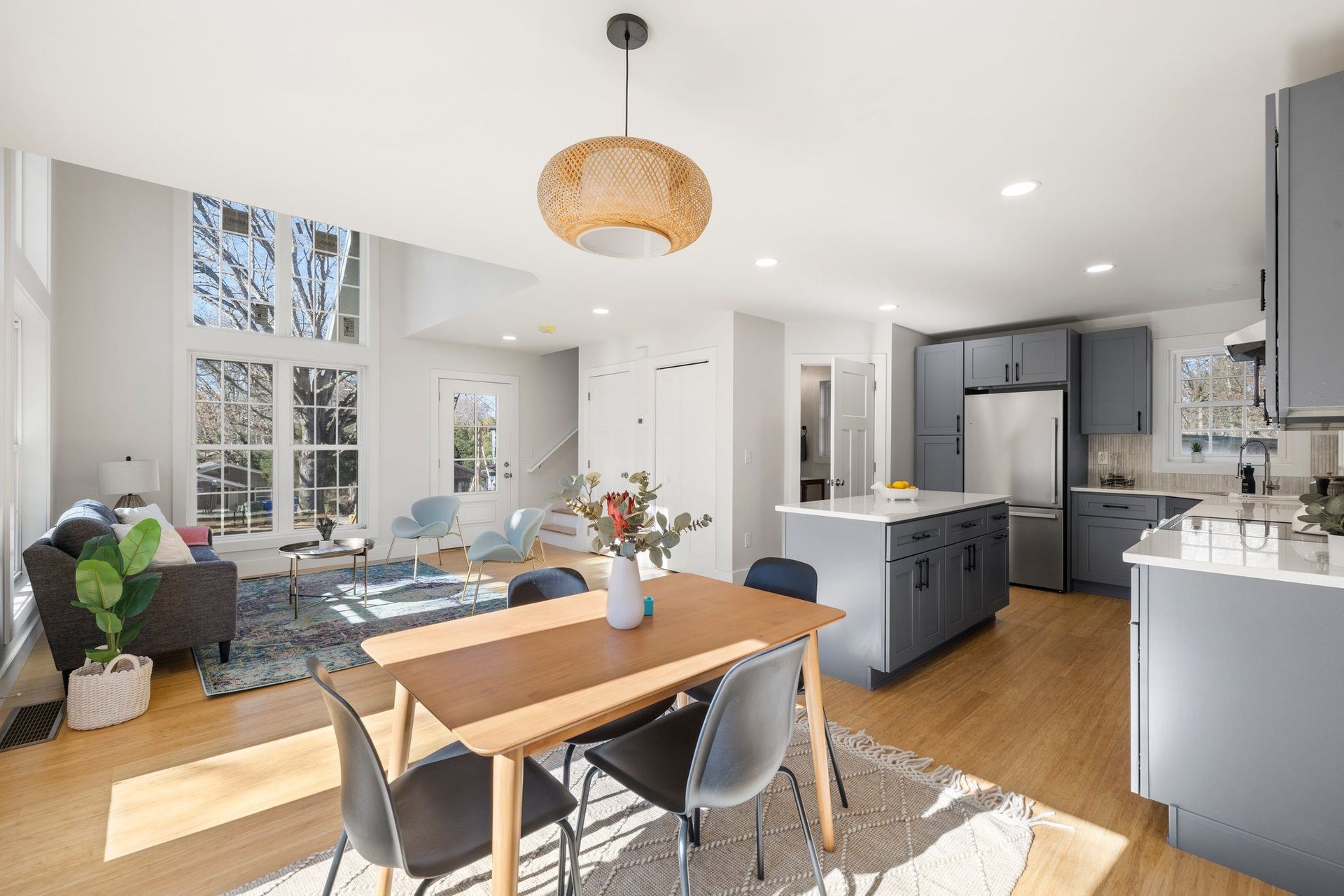 Bright open kitchen and dining area with gray cabinets, wood floors, and a table by large windows.