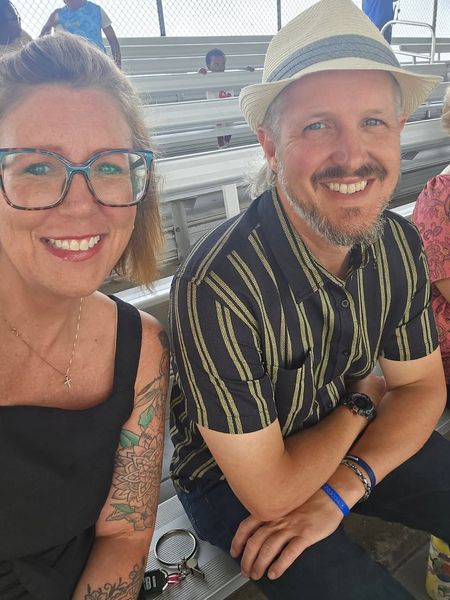 Two smiling people seated outdoors, one wearing a straw hat and striped shirt, the other glasses and a black top.