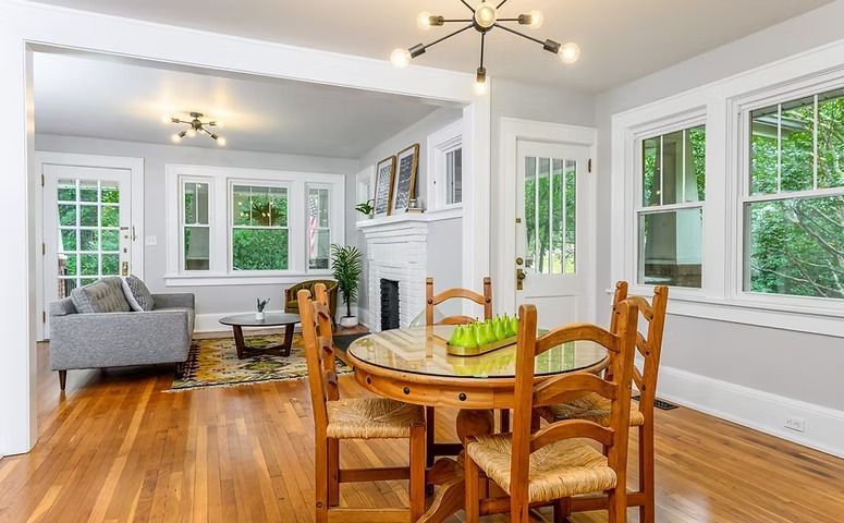 Bright dining room with wooden table, chairs, gray sofa, white trim, and large windows.