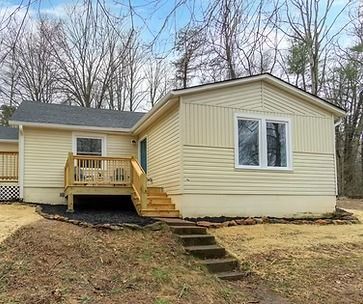 Small beige house with front porch, steps, and large window on a sloped yard