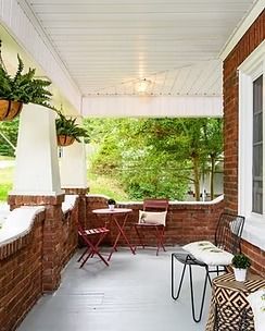Covered porch with brick walls, white ceiling, wicker chairs, and a small table overlooking green trees.