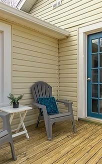 Small patio with beige siding, wooden deck, white side table, chair, and blue door with glass panels