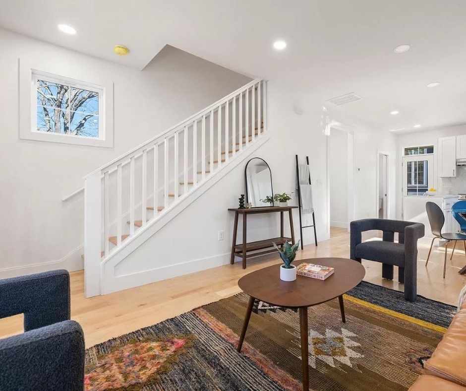 Bright living room with staircase, small table, chairs, and patterned rug in a white interior