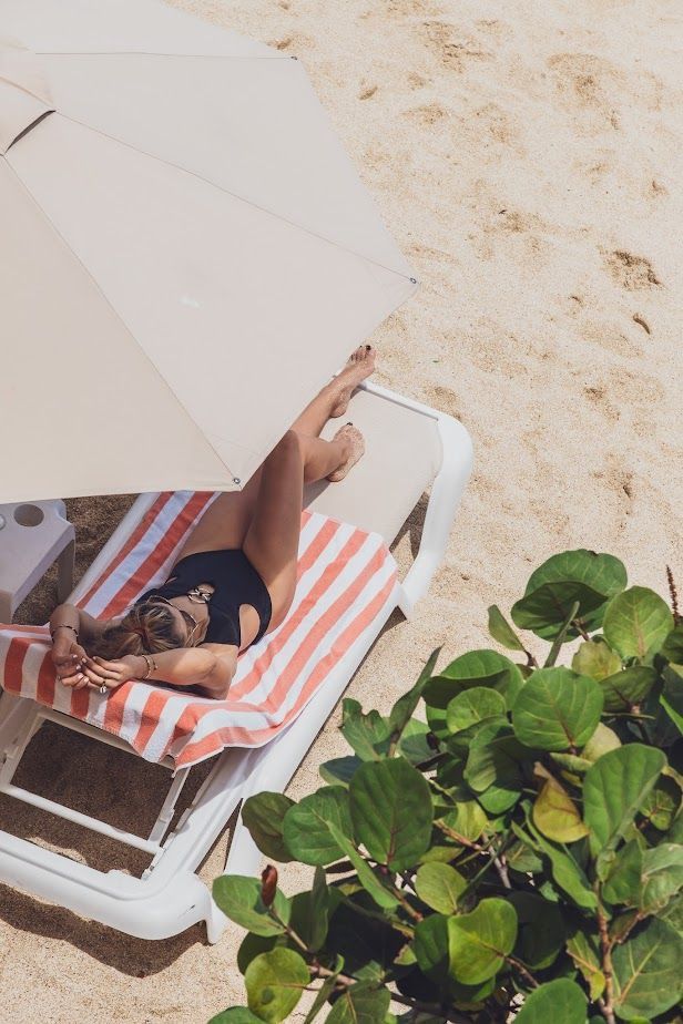 Woman in black swimsuit sunbathing on beach chair under umbrella.