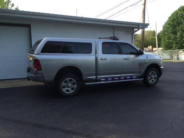 Silver pickup truck with a cap parked in front of a building. It has running board lights.