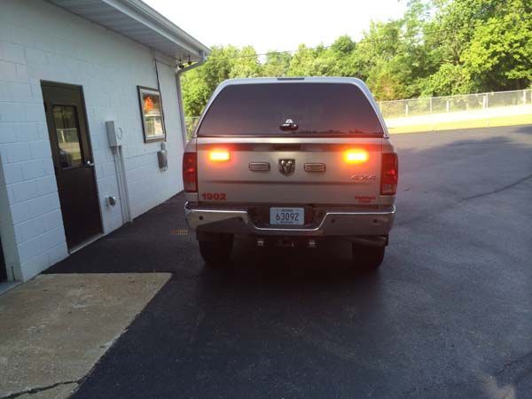 Tan Dodge pickup truck with a camper shell parked next to a white building. Tail lights illuminated.