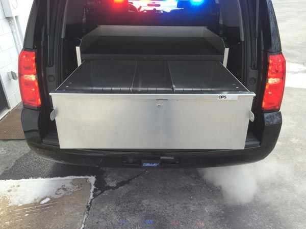 Back view of a black SUV with the trunk open, revealing a large metal storage unit inside. Police lights on.