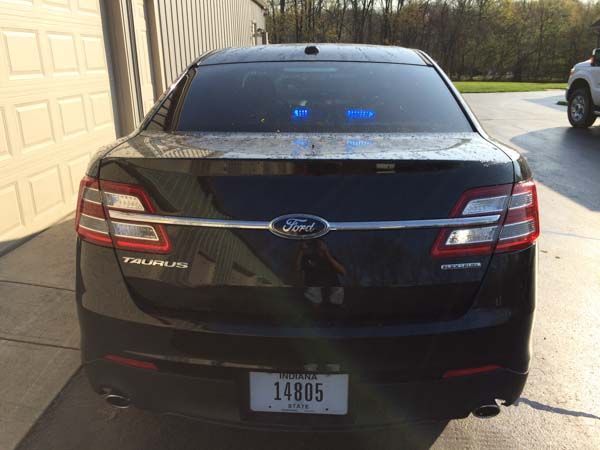 Black Ford Taurus sedan with blue flashing lights on rear window. License plate reads 
