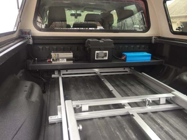 Inside truck bed: Shelf with battery and inverter, metal sliding frame on floor.